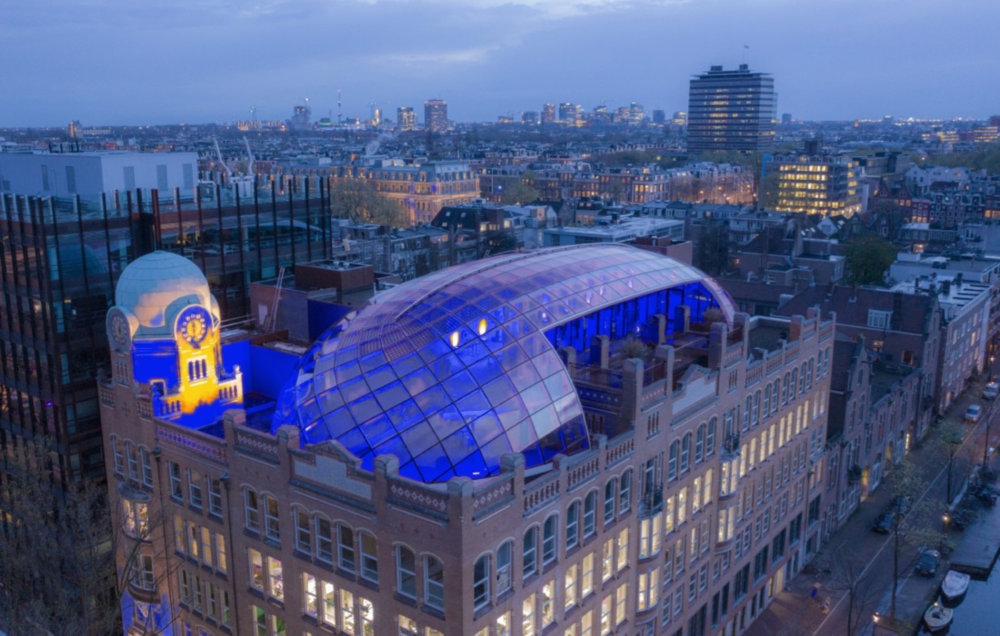 Capital C’s glass dome lit in ultramarine above the Amsterdam rooftops